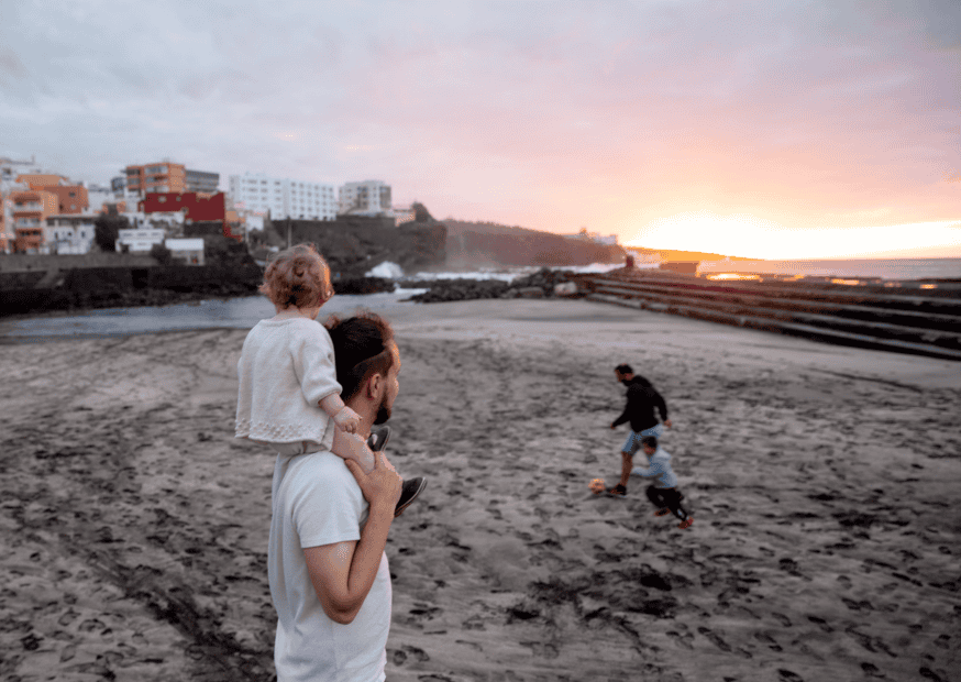 manny at the beach with child on shoulders watching people play soccer.png